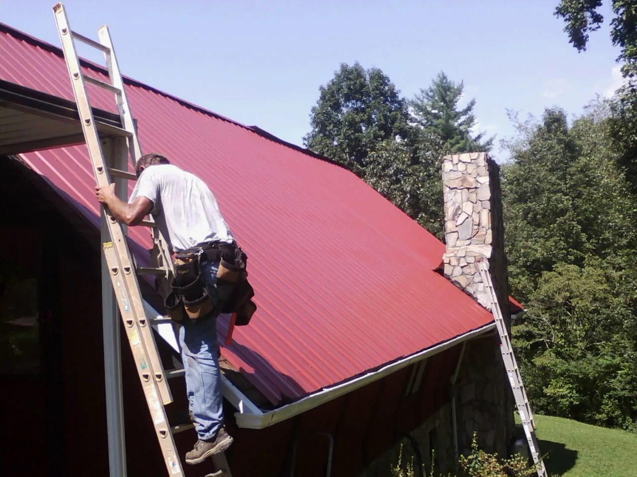 Roofer on a ladder beside a red metal roof and stone chimney — Soffit & Fascia Repair by Roofing Brothers in Bloomville
